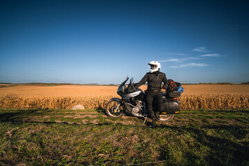 A man, a motorcycle driver, stands next to his touring motorcycle. Large side bags for luggage. A field of dry yellowed corn. Freedom and travel transportation concept.