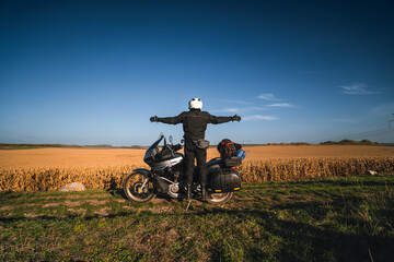 the motorcyclist stands with his arms spread out to the sides, touring motorcycle. Large side bags for luggage. A field of dry yellowed corn. Freedom and travel transportation concept.