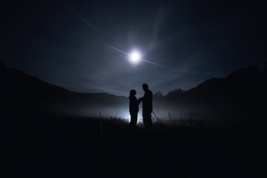 Silhouette of lovers. A couple of people, a man and a woman, are standing in the middle of a thick fog that is creeping over the ground. Mist is mystical in the moonlight. Field night
