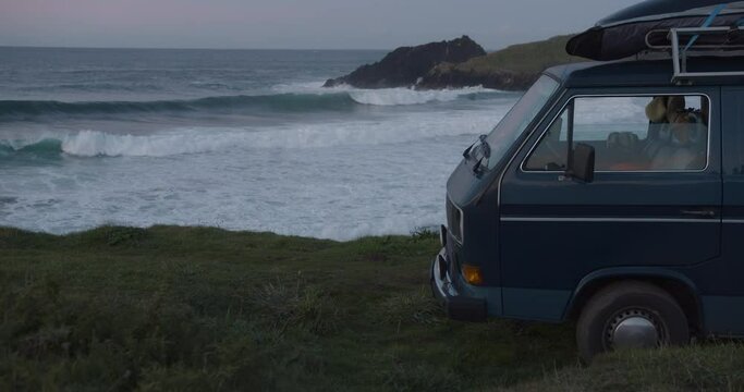 Camper van parking at the cliffs in a lonely bay during blue hour after sunset while big waves are rolling in the background in Galicia, Spain