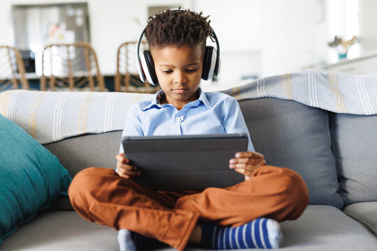 Happy african american boy sitting on sofa, using tablet in living room