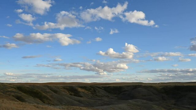 Time lapse of white clouds moving through a blue sky above a badland prairie setting with the sunlight moving on the hills.
