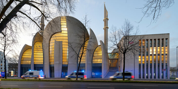 Cologne, Germany, January 11, 2023:  fast police cars with with motion blur and blue lights flashing at illuminated cologne central mosque at dusk