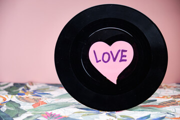 Black vinyl record with a heart in the centre above a table with a tropical pattern and a pink background.