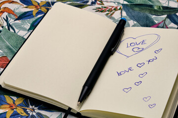 Open diary with a pen and written the word Love, on top of a table with a tropical pattern.