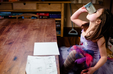 A little girl in costume sits at table sipping from mug and drawing