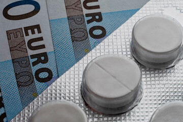 An image of a white tablet blister lying atop a set of Euro banknotes, representing the high cost and financial burden of healthcare and medication - 561143357