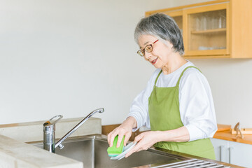家のキッチンでお皿洗いをするシニア・高齢者女性（笑顔）