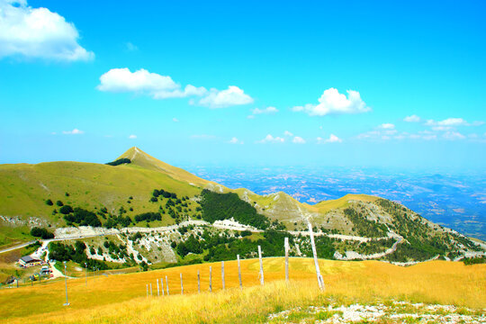 Impressive scenery from Sassotetto with yellow grass gently sitting on the slope and some majestic cusps of the Sibillini mountains interspersed with vegetation and a curvy road in the background