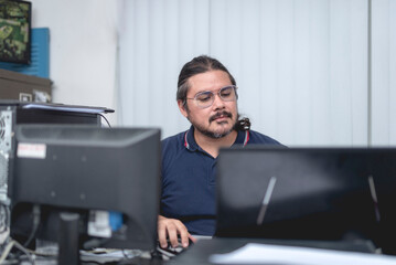 A manager in his late 30s wearing glasses and sporting tied long hair. Working at his desk checking spreadsheet data on the computer.
