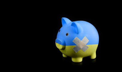 A piggy bank with a yellow-blue coloring in the style of the national flag of Ukraine and with an adhesive plaster on an imaginary wound on its side stands on a black background