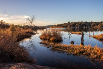 Golden hour sunset over a lakeshore changing the colors of the trees and grass to yellow and orange. Many people visit Inks Lake State Park in Texas during the Fall and early Spring