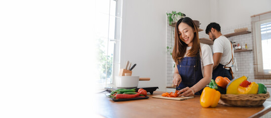 Happy asia young couple cooking together with vegetables in cozy kitchen, preparing vegetarian food colorful variety of vegetables and herbs lying on wooden kitchen table, love and valentine concept.
