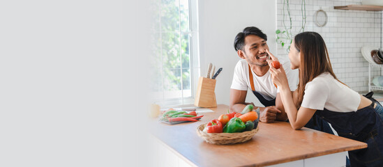 Happy asia young couple cooking together with vegetables in cozy kitchen, preparing vegetarian food colorful variety of vegetables and herbs lying on wooden kitchen table, love and valentine concept.