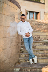 Obraz premium Handsome man in a trendy white shirt and sunglasses in the city. Handsome and charming man posing outdoors, urban style
