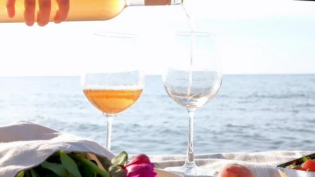 Hand pouring rose wine into glasses with a calm sea on the background. Summer picnic