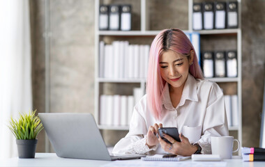 Fototapeta premium Portrait of a happy Asian businesswoman working on a laptop computer at her home office while using a mobile phone. Smiling businesswoman contacting the business to talk about online products.