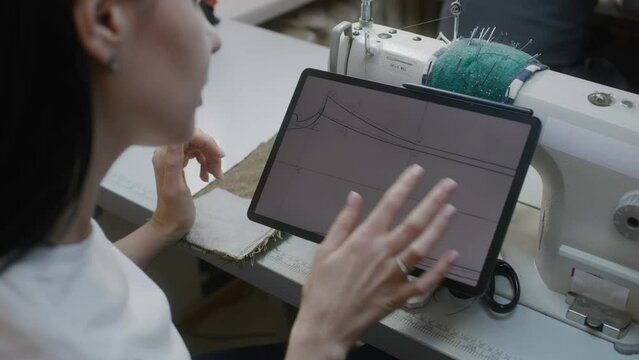 Female tailor sits at sewing machine with digital tablet and zooming picture. Dressmaker or seamstress watches sizes of custom apparel at sketch and sewing suit. Atelier workshop. Concept of fashion.