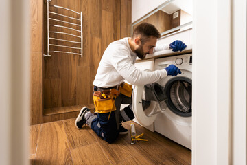 Plumber in overalls with tools is repairing a washing machine in the house.