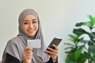 Millennial Arab woman smiling while holding a credit card and smartphone, Shopping Online, Using Credit Card Making Payment, Internet Banking Application and E-Commerce concept.