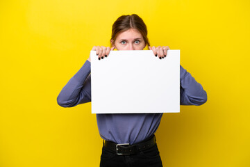 Young English woman isolated on yellow background holding an empty placard and hiding behind it
