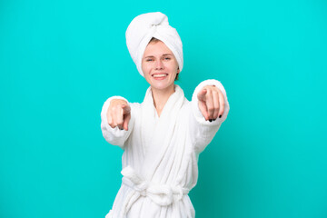 Young woman in a bathrobe isolated on blue background points finger at you while smiling