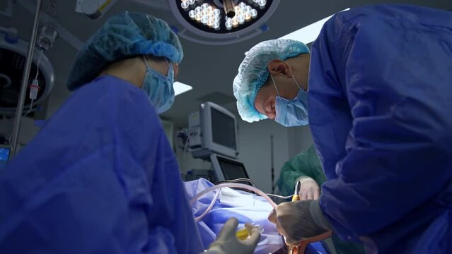 Male Caucasian surgeon uses tools in both hands at operation. Nurse holds the syringe ready to assist the doctor. Low angle view.