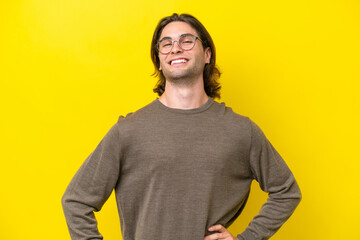 Caucasian handsome man isolated on yellow background posing with arms at hip and smiling