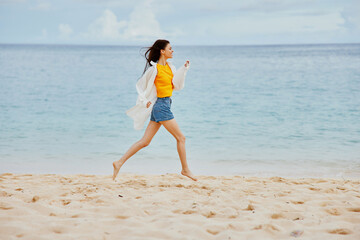 Fototapeta na wymiar Sports woman runs along the beach in summer clothes on the sand in a yellow T-shirt and denim shorts white shirt flying hair ocean view