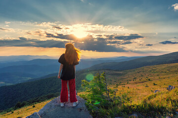 Naklejka premium A young, beautiful girl with flowing red hair against the backdrop of a sunset in the mountains. Beauty stands on a rock