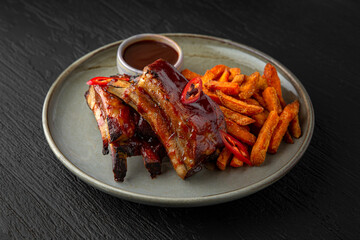 Pork ribs with sweet potato fries in a ceramic plate on a dark textured background. Restaurant menu Isolated on black