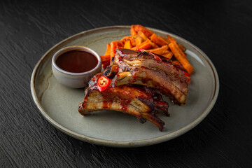 Pork ribs with sweet potato fries in a ceramic plate on a dark textured background. Restaurant menu Isolated on black