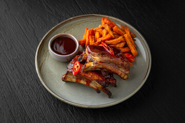 Pork ribs with sweet potato fries in a ceramic plate on a dark textured background. Restaurant menu Isolated on black