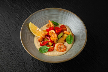 Hummus with vegetables and shrimp in a ceramic plate on a dark textured background. Restaurant menu Isolated on black