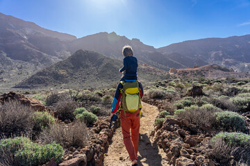 Aktiv auf Teneriffa, Kanarische Inseln: Wanderung im Teide Gebiet zum dritthöchsten Gipfel Alto de Guajara - Mann mit Kind auf dem Rücken wandert in der Sonne - obrazy, fototapety, plakaty