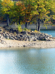 Beautiful scenery of colorful trees on a rugged rock, sunlit on a late summer day. The surrounding lake reflects the bright blue sky.