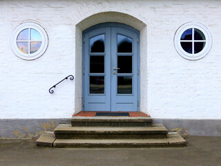 Entrance to an old public building. In front of the steel-blue doors are three steps, and on the left and right side of the door are two round windows.