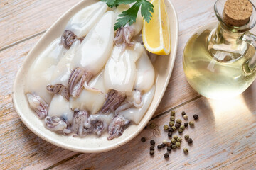 Raw squid fillet on a plate over wooden table. Small calamary tubes, olive oil and lemon prepared for cooking. Fresh squid ingredient for low calorie diet. Seafood concept.