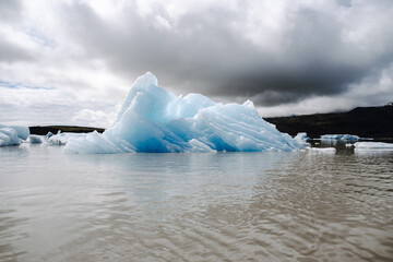 View of a blue iceberg broken off from the glacier tongue of Fjallsj&ouml;kull in the glacier lagoon Fjalls&aacute;rl&oacute;n in Iceland on a cloudy day. With a view of &Ouml;r&aelig;faj&ouml;kull in the background.