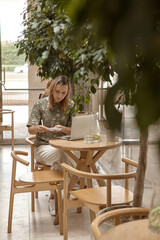 A young woman is sitting in a cafe and working on a laptop. The concept of freelancing and remote work or training. A woman makes notes in a notebook and looks at the laptop screen