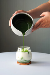 Preparing matcha latte green tea with milk in a glass on white table over gray background, hand hold a bowl pouring matcha to the glass with ice cubes.