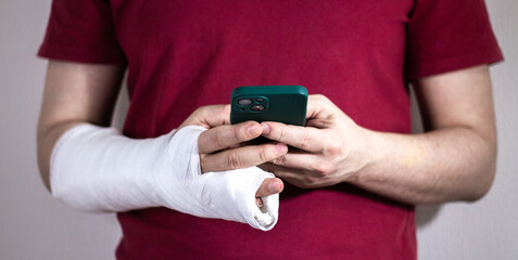 A man in a red T-shirt with a broken arm in a cast holds and uses a phone Close-up