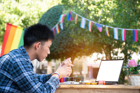 Young asian teen gay in plaid shirt wears rainbow wristband, holds brush and drawing rainbow heart in the park which decorated with LGBTQ+ flaglines, concept for hobby of LGBT people over the world.