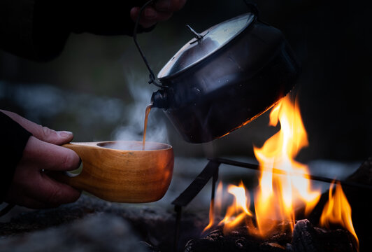 Hands pouring coffee from a cettle into a wooden cup at a campfire
