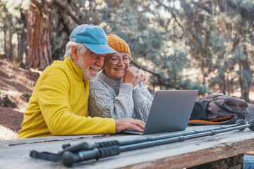 Head shot portrait close up of cute couple of old middle age people using computer pc outdoors sitting at a wooden table in the forest of mountain in nature with trees around them.