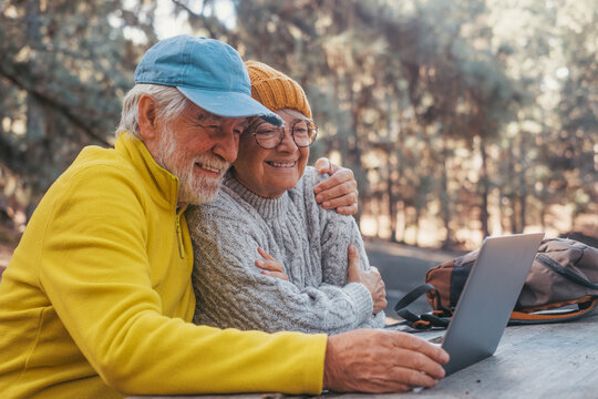 Head shot portrait close up of cute couple of old middle age people using computer pc outdoors sitting at a wooden table in the forest of mountain in nature with trees around them.