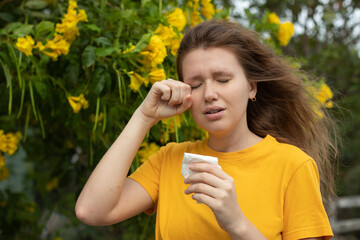 Fototapeta premium Portrait of beautiful young allergic woman is suffering from pollen allergy or cold on natural flower flowering tree background at spring or sunny summer day sneezes, blowing her runny nose rubs eyes