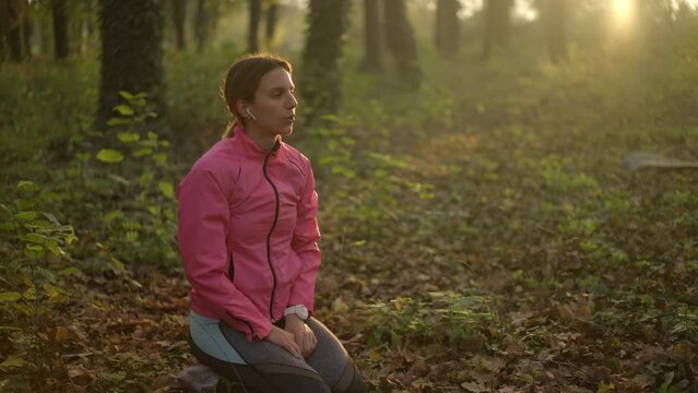 Relaxed woman doing breathing exercise and listening music with headphones on the yoga mat in the forest