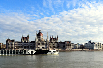 Fototapeta premium the Hungarian Parliament in Neo-Gothic style from across the Danube in panoramic view. white tour and hotel boat going by. blue sky. Famous and popular Budapest landmark and attraction. City skyline.