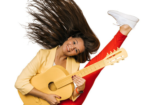 Young attractive brunette girl dressed in yellow jacket and red pants holding yellow guitar dancing with fluttering long hair and raising her leg high on transparent background. Happy people concept.